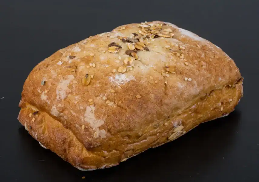 A freshly baked ciabatta bread with a golden crust, topped with sesame seeds, oats, and grains, placed on a black background.