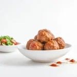 A white bowl filled with Italian meatballs in a light tomato sauce, placed on a white surface with a side bowl of fresh tomato and arugula salad. Small drops of tomato sauce and dried herbs are scattered nearby.