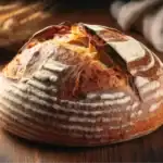 A beautifully baked sourdough bread loaf with a deep golden crust, flour-dusted surface, and rustic scoring, placed on a wooden background.