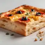 Thick slice of golden Italian focaccia bread topped with cherry tomatoes, black olives, and herbs.