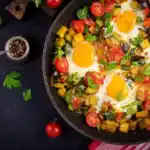 Colorful shakshuka with eggs, cherry tomatoes, zucchini, and herbs in a black skillet