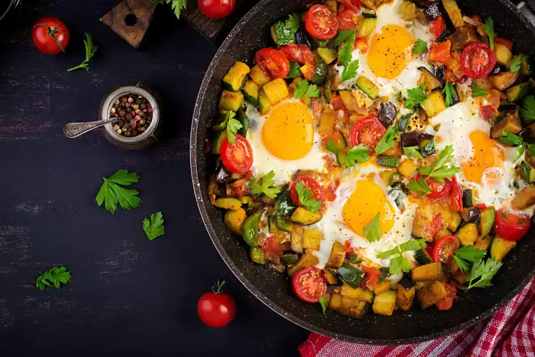 Colorful shakshuka with eggs, cherry tomatoes, zucchini, and herbs in a black skillet