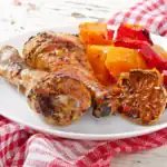 Roasted chicken drumstick served with garlic, sweet potatoes, and red bell peppers on a white plate.