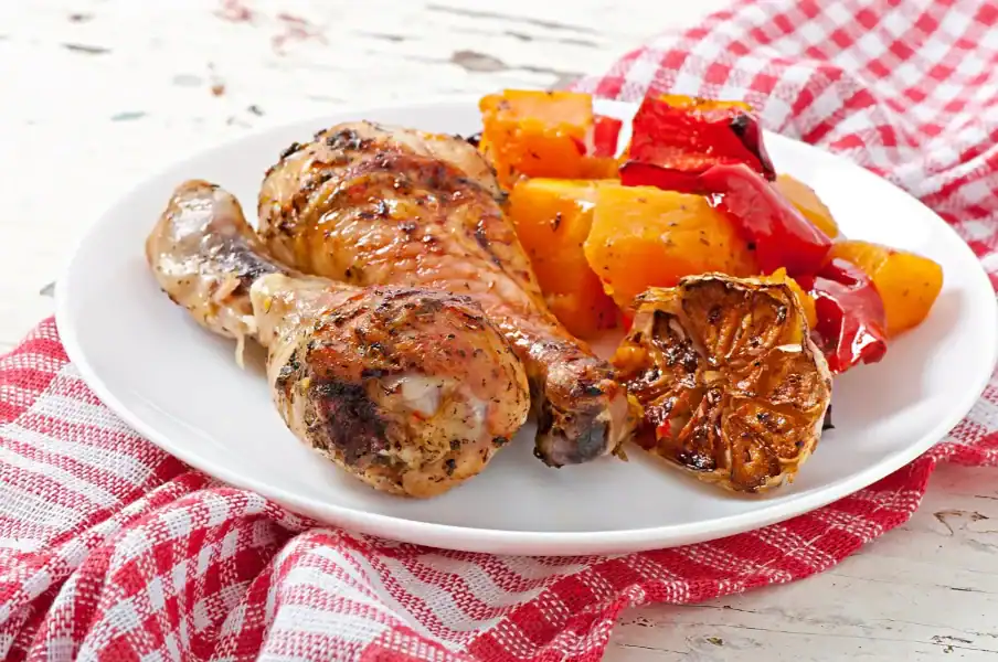 Roasted chicken drumstick served with garlic, sweet potatoes, and red bell peppers on a white plate.