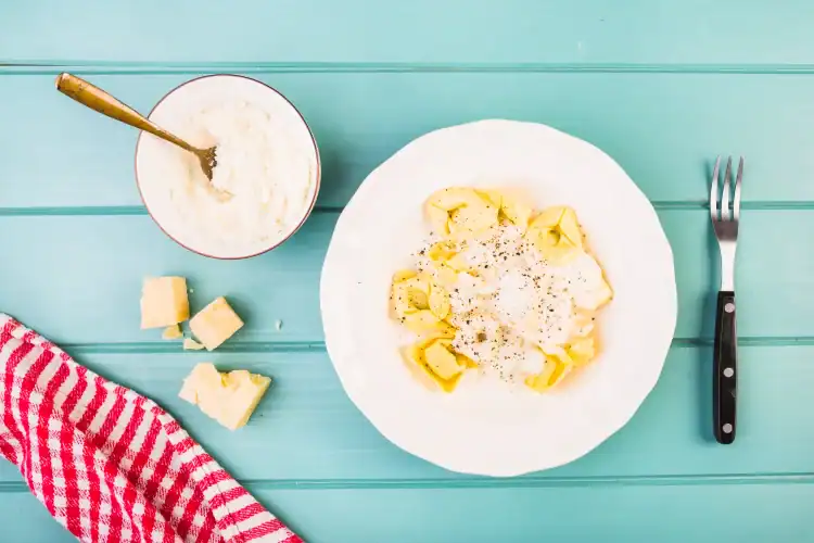 Plate of ricotta-filled pasta topped with Parmesan cheese and black pepper, served with a bowl of grated cheese and Parmesan chunks on a turquoise table.