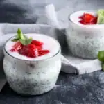 Two glass jars of chia pudding topped with strawberry slices and mint leaves, placed on a gray surface with a light cloth and fresh mint nearby