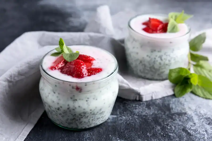 Two glass jars of chia pudding topped with strawberry slices and mint leaves, placed on a gray surface with a light cloth and fresh mint nearby