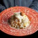 Hand holding a vibrant orange plate of creamy ricotta pasta topped with Parmesan cheese and an artistic garnish.