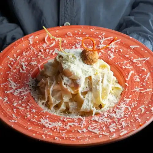 Hand holding a vibrant orange plate of creamy ricotta pasta topped with Parmesan cheese and an artistic garnish.