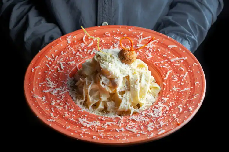 Hand holding a vibrant orange plate of creamy ricotta pasta topped with Parmesan cheese and an artistic garnish.