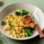 Juicy baked chicken thigh served with seasoned rice, broccoli, and fresh herbs on a plate.