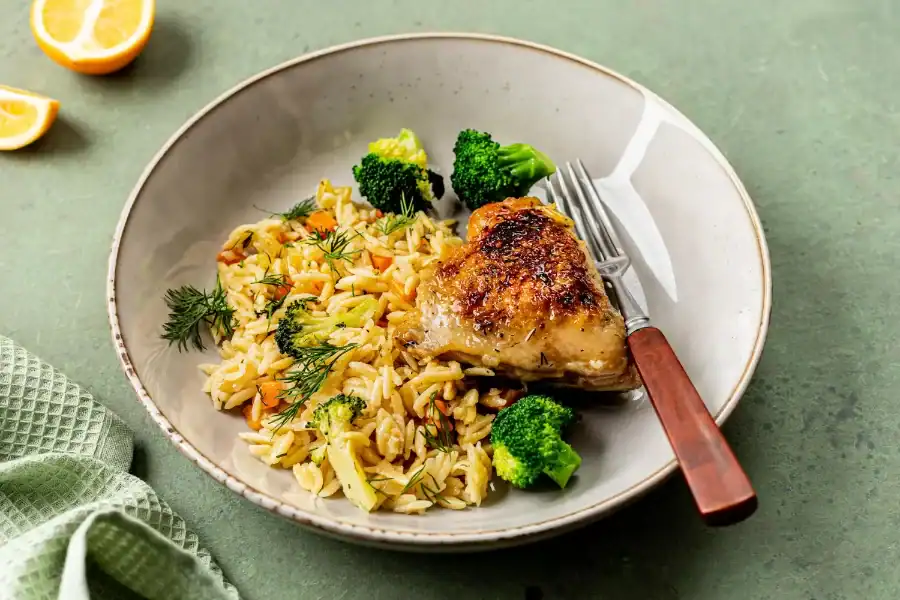 Juicy baked chicken thigh served with seasoned rice, broccoli, and fresh herbs on a plate.