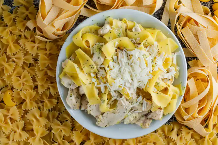 Plate of creamy pasta with chicken and Parmesan cheese, surrounded by uncooked fettuccine and farfalle pasta.