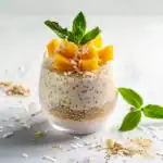 A glass of layered coconut chia pudding topped with fresh mango chunks, shredded coconut, and mint leaves.