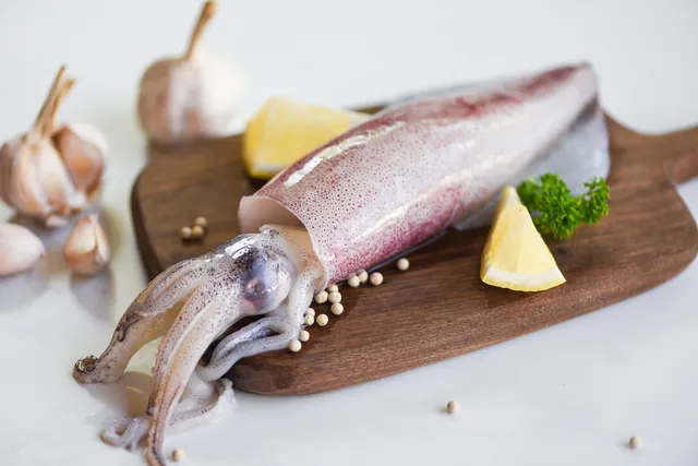 Raw whole squid on a wooden board with garlic, lemon wedges, and peppercorns