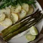 Roasted asparagus spears served with sliced potatoes, arugula, lime wedges, and a creamy sauce on a white plate.