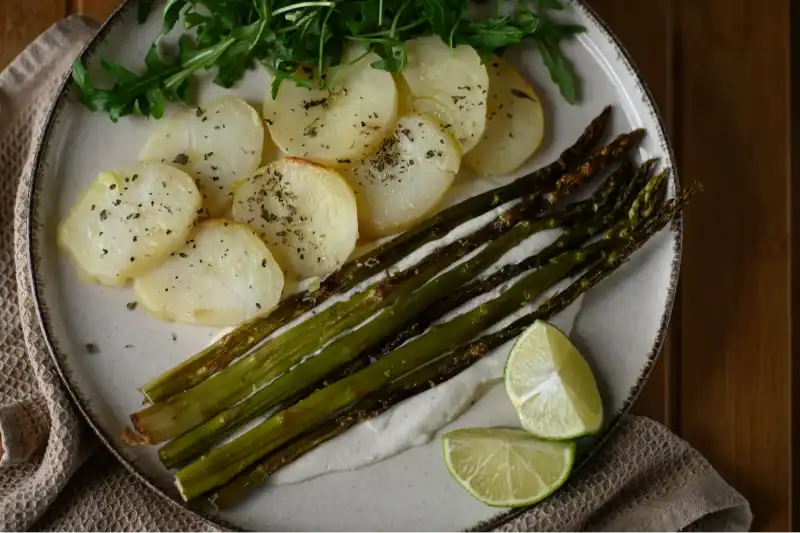 Roasted asparagus spears served with sliced potatoes, arugula, lime wedges, and a creamy sauce on a white plate.