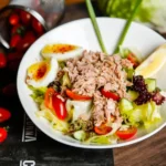 Tuna salad in a white bowl with cherry tomatoes, soft-boiled eggs, cucumber, lettuce, and a lemon wedge