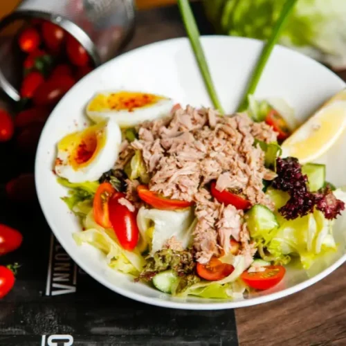 Tuna salad in a white bowl with cherry tomatoes, soft-boiled eggs, cucumber, lettuce, and a lemon wedge