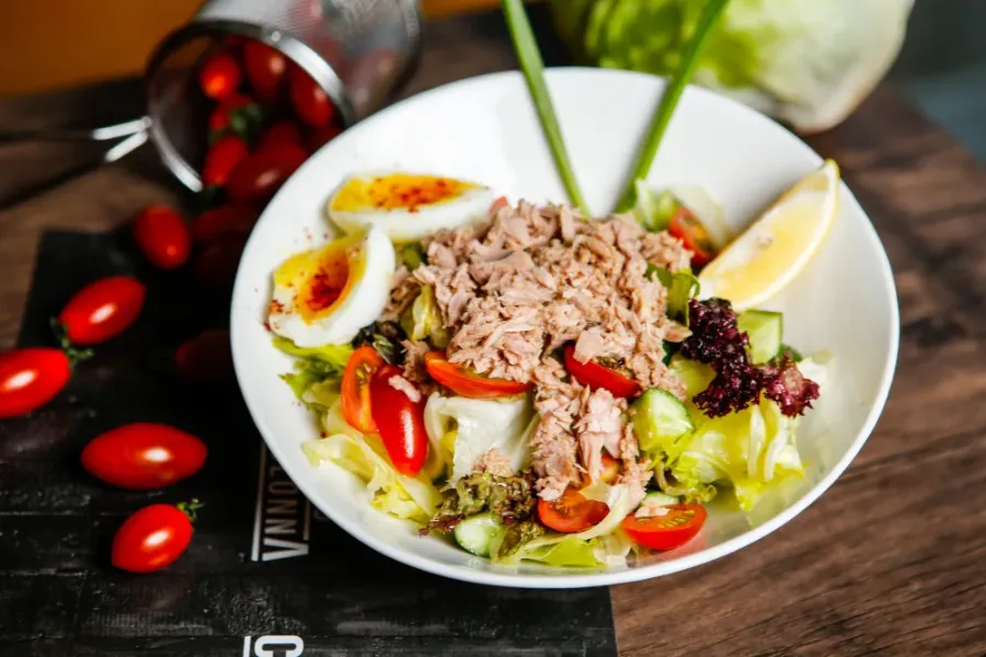 Tuna salad in a white bowl with cherry tomatoes, soft-boiled eggs, cucumber, lettuce, and a lemon wedge