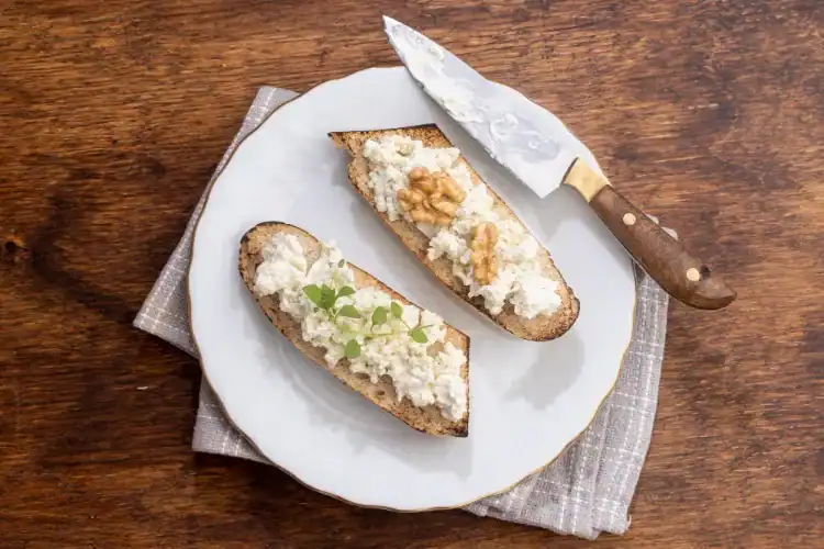 Two slices of toasted bread topped with ricotta cheese, walnuts, and herbs on a white plate.