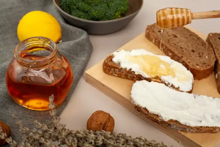 Slices of whole grain bread topped with ricotta cheese and honey, surrounded by honey jar, lemon, and nuts.