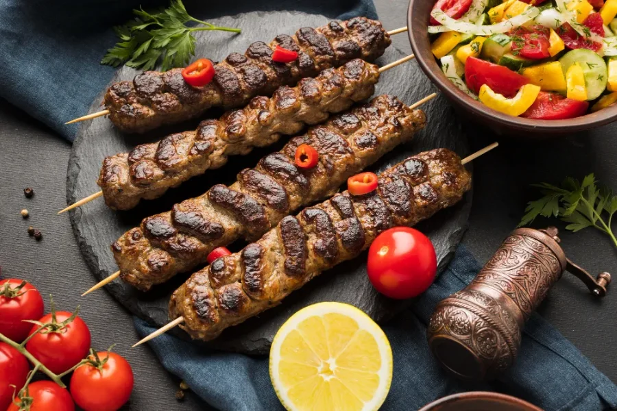 Grilled kofta kebabs on skewers served with fresh tomatoes, lemon, and mixed vegetable salad