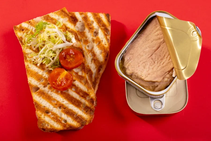 Grilled sandwich bread with shredded lettuce and cherry tomatoes next to an open can of tuna on a red background