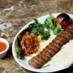 Grilled Adana kebab on a plate with flatbread, grilled vegetables, parsley, and spicy sauce