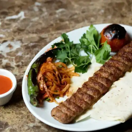Grilled Adana kebab on a plate with flatbread, grilled vegetables, parsley, and spicy sauce