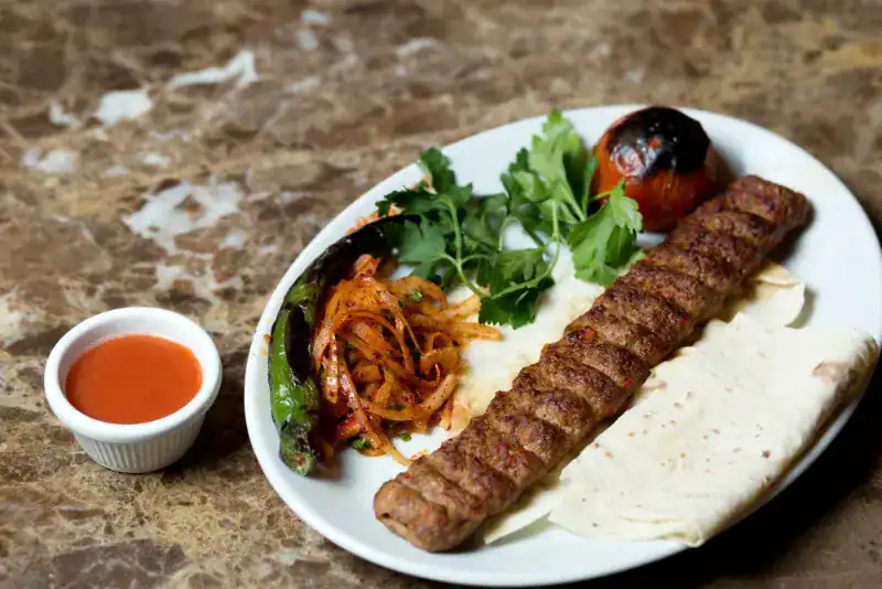 Grilled Adana kebab on a plate with flatbread, grilled vegetables, parsley, and spicy sauce