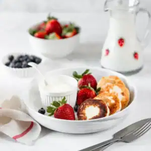 Ricotta pancakes served with fresh strawberries, blueberries, and a side of yogurt in a white bowl on a breakfast table.