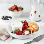 Ricotta pancakes served with fresh strawberries, blueberries, and a side of yogurt in a white bowl on a breakfast table.