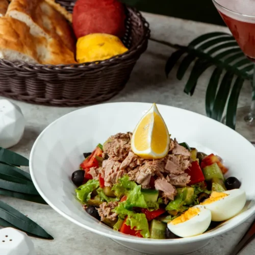 Tuna salad served in a white bowl with lettuce, boiled egg, vegetables, olives, and a lemon wedge