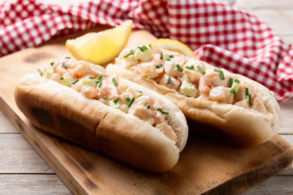 Two homemade Maine-style lobster rolls with creamy lobster filling, chopped chives, and lemon wedges on a wooden board.
