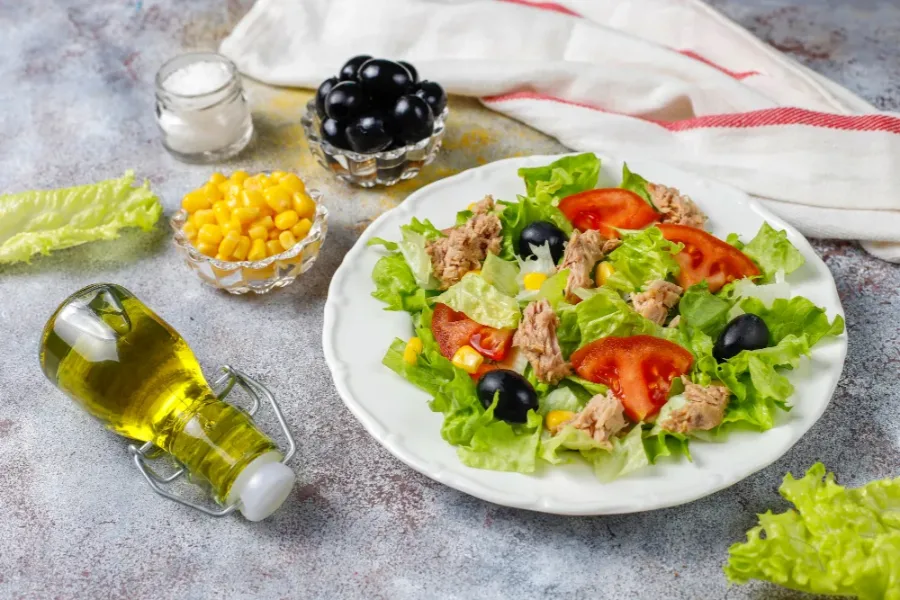 Tuna salad with lettuce, tomato, corn, and black olives on a white plate, surrounded by olive oil, salt, and ingredients