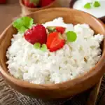 Fresh ricotta cheese in a wooden bowl topped with strawberries and mint leaves on a rustic wooden table.