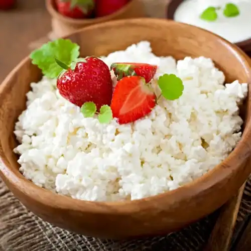 Fresh ricotta cheese in a wooden bowl topped with strawberries and mint leaves on a rustic wooden table.
