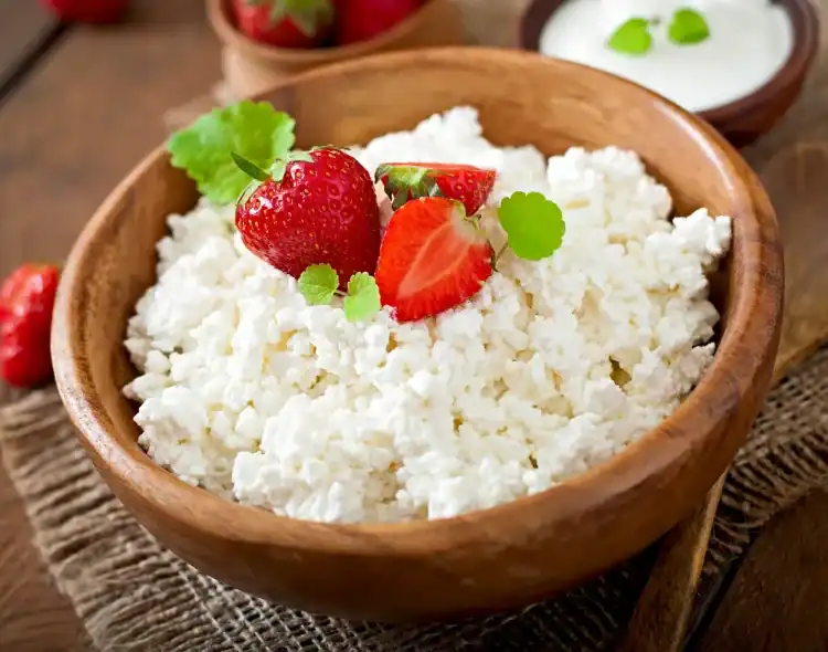 Fresh ricotta cheese in a wooden bowl topped with strawberries and mint leaves on a rustic wooden table.