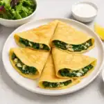 Four spinach and cheese crepes served with salad, yogurt sauce, lemon wedge, and cucumber slices on a white plate.