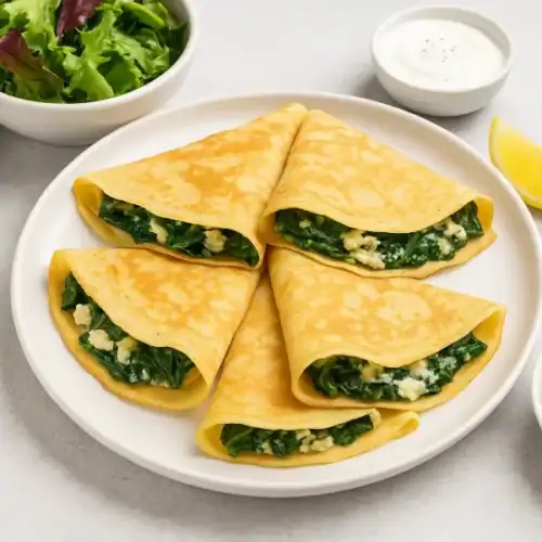 Four spinach and cheese crepes served with salad, yogurt sauce, lemon wedge, and cucumber slices on a white plate.