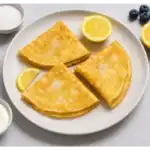 Three folded lemon and sugar crepes on a white plate, served with lemon wedges, blueberries, and yogurt on a light grey background.