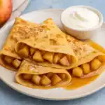 Three golden apple cinnamon crepes filled with soft spiced apples, served with syrup and whipped cream on a white plate.