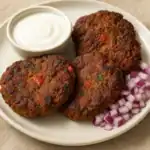 Four chapli kebabs served with yogurt sauce and diced red onions on a white plate