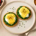 Eggs Florentine served on a plate with hollandaise sauce, spinach, poached eggs, and brunch sides like fruit, potatoes, and orange juice.