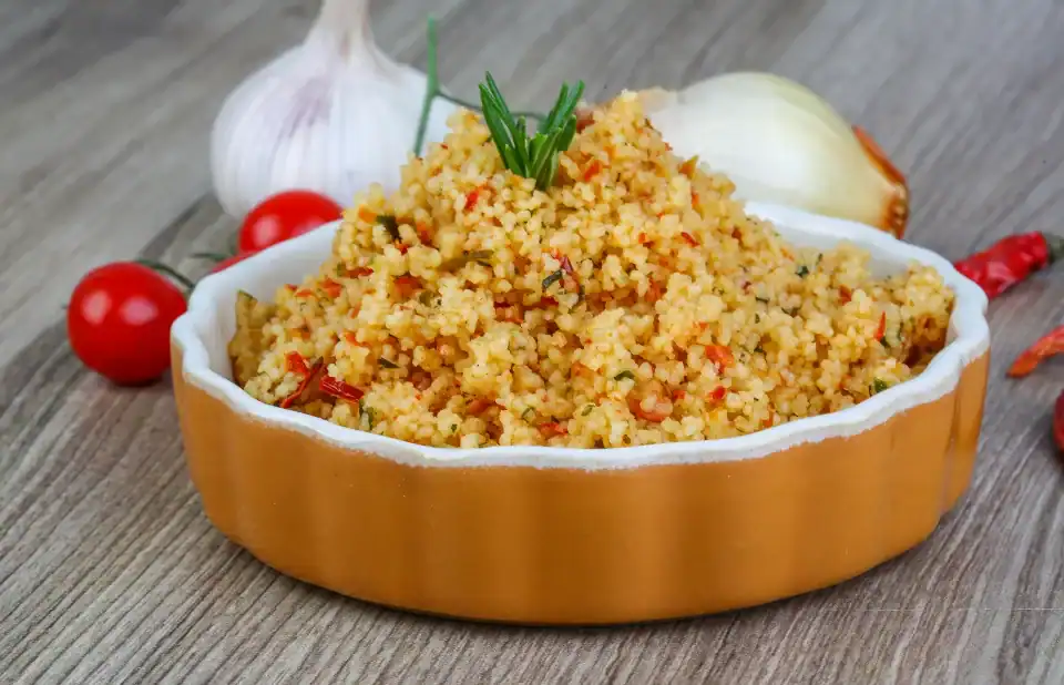 A bowl of seasoned couscous garnished with herbs, surrounded by fresh garlic, cherry tomatoes, and onion on a wooden surface.