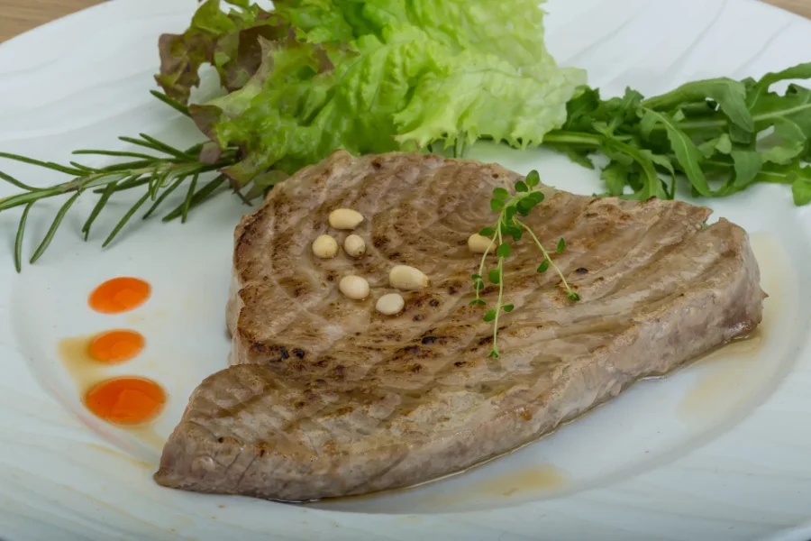 Medium-grilled tuna steak garnished with pine nuts, fresh herbs, and served with leafy greens.