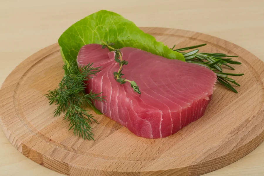 Raw tuna steak on a wooden board, garnished with rosemary, dill, and lettuce leaves.