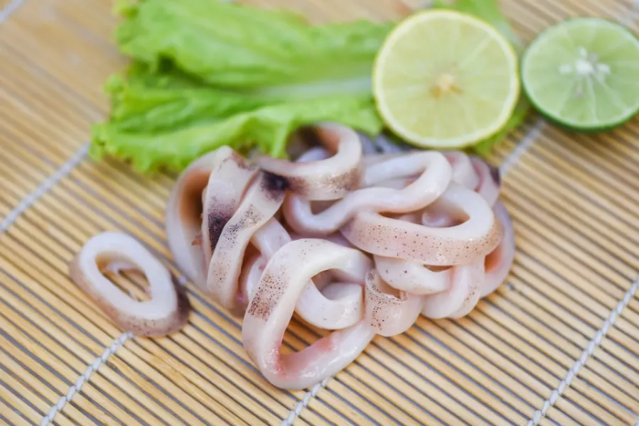 Raw squid rings prepared for cooking, placed on a bamboo mat with lettuce, lemon, and lime.