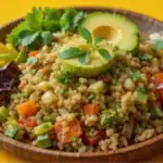 Couscous salad with avocado, cherry tomatoes, broccoli, and fresh herbs in a wooden bowl on a yellow background.